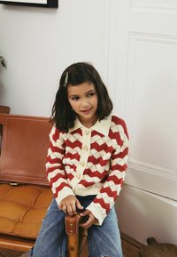 Cardigan en tricot à motifs rouges et crème avec un col et des boutons, associé à un jean bleu, sur fond neutre.