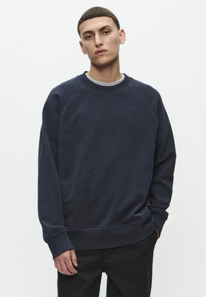 Mladý muž s krátkymi vlasmi, oblečený v tmavomodrej mikine cez sivé tričko, stojaci so uvoľnenými rukami, neutrálneho výrazu, na bielom pozadí.