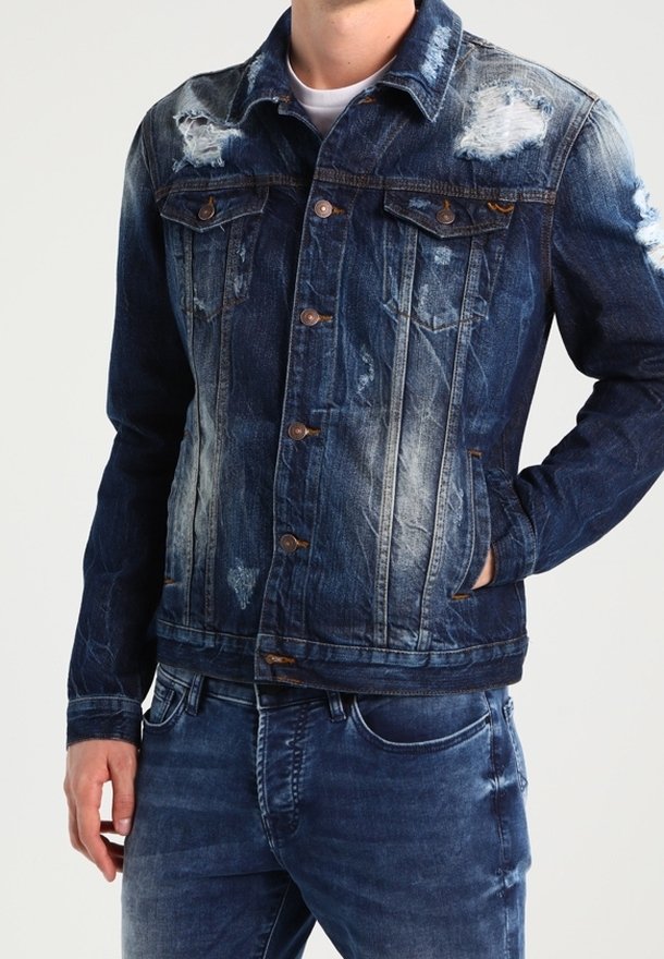 Man med en sliten blå denimjacka med knäppning och matchande blå jeans, med en hand i fickan.