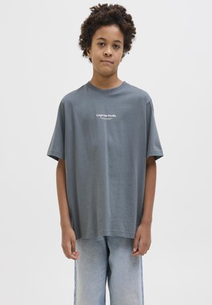 Garçon aux cheveux bouclés portant un t-shirt gris ample avec le texte "Originals Studio" et un jean bleu clair, debout devant un fond uni.