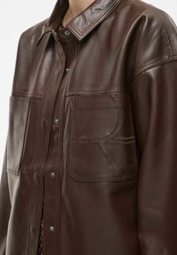 Veste en cuir marron avec un col, deux poches avant et des fermetures à pression. Texture lisse avec des détails de coutures visibles.