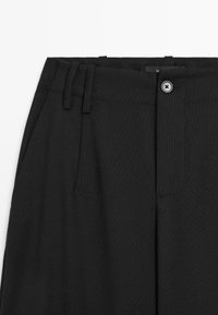 Zwarte broek met een gladde textuur, plooien en een knoopsluiting. Inclusief riemlussen en een gestructureerd ontwerp van de tailleband.