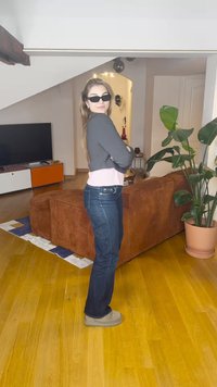 Egy modell szürke, rövidített pulóvert visel rózsaszín felső fölött, sötét farmert és világosbarna cipőt, miközben egy barna kordbársony kanapé mellett áll egy nappaliban.