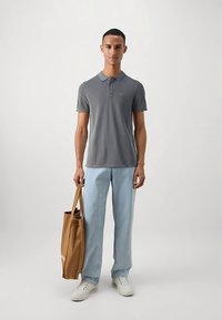 Sivá polokošeľa s golierom, svetlomodré rifle a biele tenisky, v ruke hnedá taška na plece. Jednoduchý, ležérny outfit s neutrálnou farebnou paletou.