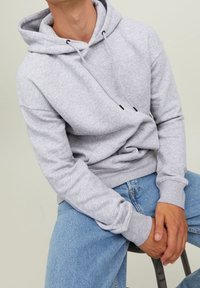 Felpa grigia con cappuccio e lacci, polsini e orlo a coste. Vestibilità comoda. Indossata sopra jeans blu, seduta su una sedia.