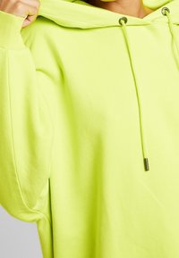 Felpa oversize neon verde realizzata in un tessuto morbido; presenta un cappuccio con cordoncino e polsini a coste, con una leggera cucitura laterale.