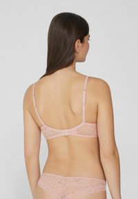 Svetle růžová krajková podprsenka s nastavitelnými ramínky a háčkovým zapínáním, zdobená květinovými vzory a hladkým povrchem. Pohled na záda modelky.