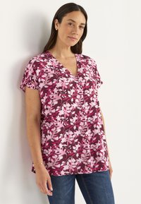 Kvinde lænet op ad væggen iført en lyserød og burgunder blomstret V-hals bluse med korte ærmer og blå jeans.