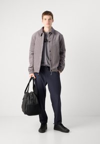 Grijze corduroy jas, grijs T-shirt, marineblauwe broek, zwarte schoenen. Model houdt een zwarte tas vast met verstelbare schouderbanden. Eenvoudig ontwerp, casual uitstraling.