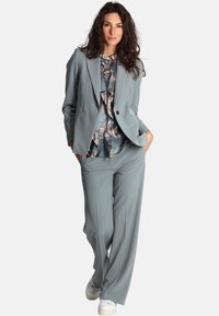 Completo grigio su misura con blazer a un bottone, pantaloni a gamba ampia e una maglietta a fantasia con motivi fogliati. Sneakers bianche semplici completano il look.