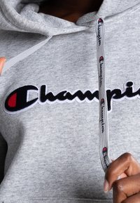 Sudadera gris hecha de un tejido suave, que presenta un destacado logo bordado en negro y rojo. Incluye un cordón gris con acentos del logo.