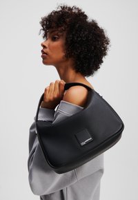 Borsa a tracolla nera in pelle con una texture liscia, forma rettangolare e una patch del logo ben evidente. Progettata con una breve tracolla.