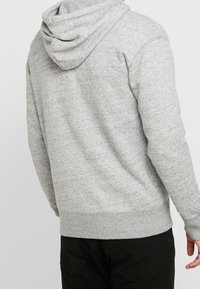 Uomo che indossa una felpa con cappuccio grigio chiaro a maniche lunghe e pantaloni neri, visto di spalle su uno sfondo semplice.