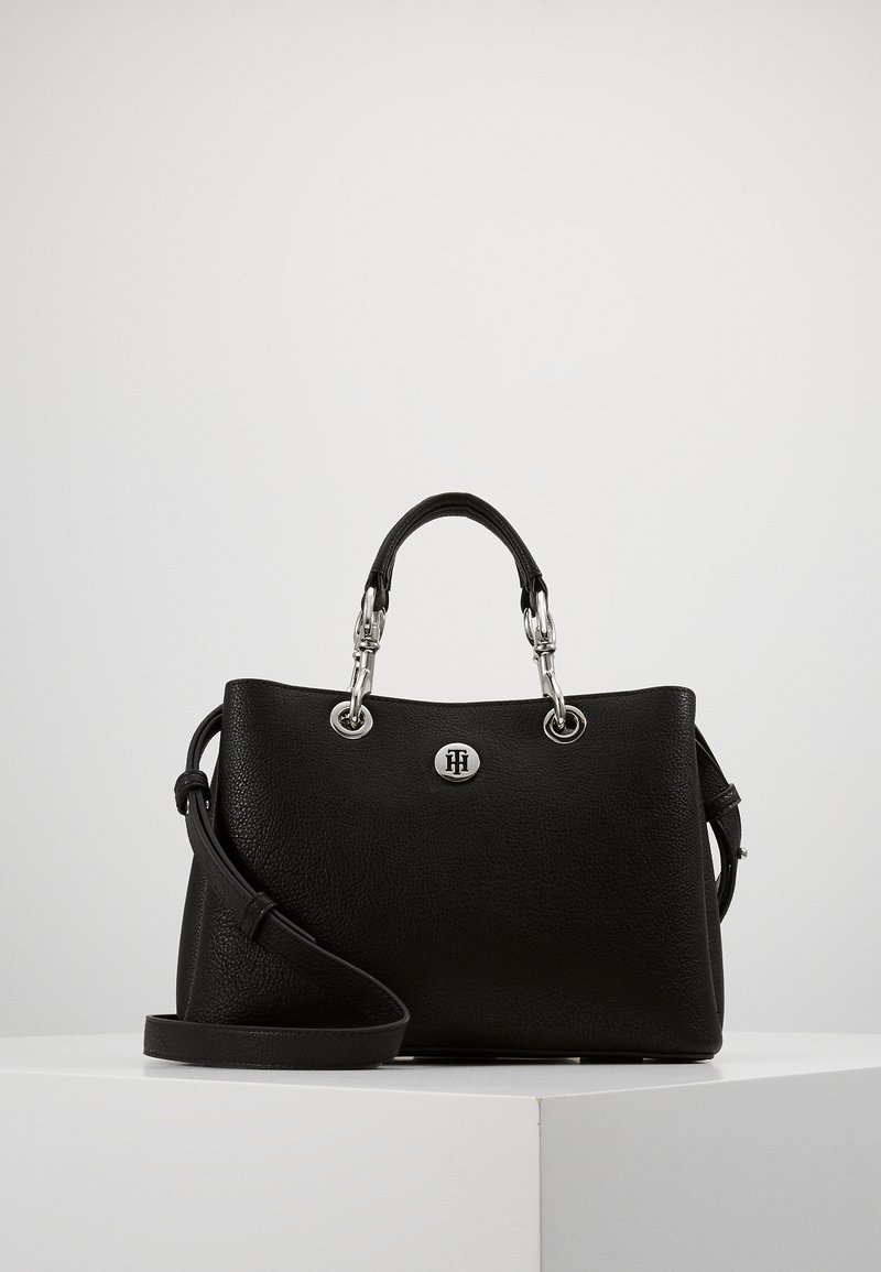 Sac à main en cuir noir avec poignées supérieures, quincaillerie argentée, logo circulaire de la marque et bandoulière amovible sur surface blanche.