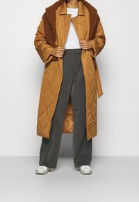 Abrigo acolchado marrón con un amplio cuello, llevado abierto sobre una blusa blanca, combinado con pantalones de pierna ancha grises y zapatos blancos.
