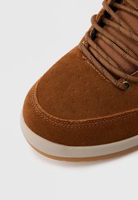 Smeđa tenisica od brušene kože s zaobljenim vrhom, mrežastim unutrašnjim slojem, teksturiranim detaljima i potplatom od smeđe gume. Ima kontrastne šavove i metalne rupice za vezice.