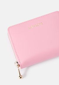 Lichtroze ritsportemonnee van glad synthetisch materiaal, met een gouden logo, een gouden rits en afgeronde hoeken.