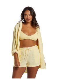 Kvinde iført en gul tekstureret bralette, matchende shorts med snørelukning og en åben gul skjorte, poserende mod hvid baggrund.