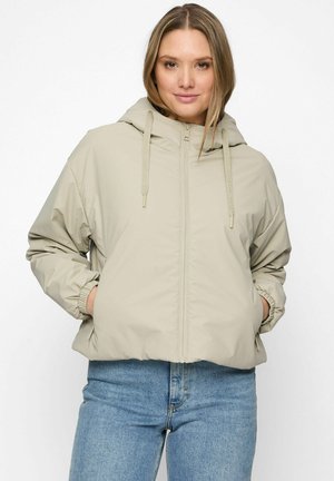 Vrouw die een beige gewatteerd jack met capuchon draagt, met handen in de zakken en een blauwe spijkerbroek, staand tegen een effen witte achtergrond.