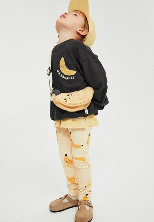 Kind met een gele pet, zwart shirt met de print "Go Bananas", broek met bananenpatroon, beige heuptasje en bruine schoenen, kijkt omhoog.