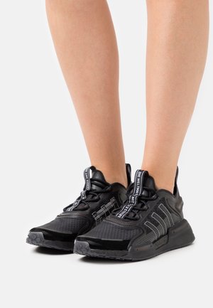 Pieds portant des baskets de sport noires en mesh texturé avec des rayures réfléchissantes, debout sur un fond blanc uni.