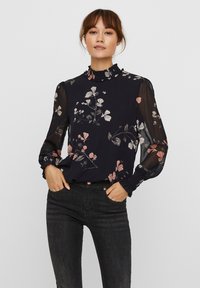 Blusa floreale navy con maniche lunghe trasparenti, scollatura a volant e motivi floreali multicolore. Abbinata a jeans in denim scuro.