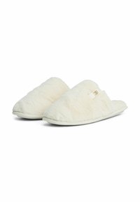 Een paar witte, pluizige instappers met platte zolen en een klein stoffen label aan de zijkant, weergegeven tegen een witte achtergrond.