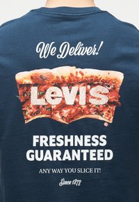 Η πίσω πλευρά της μπλούζας με γραφικό κομμάτι πίτσας που σχηματίζει τη λέξη «Levi's», με το κείμενο πάνω να λέει «We Deliver!» και κάτω «Freshness Guaranteed, Any way you slice it! Since 1873.»