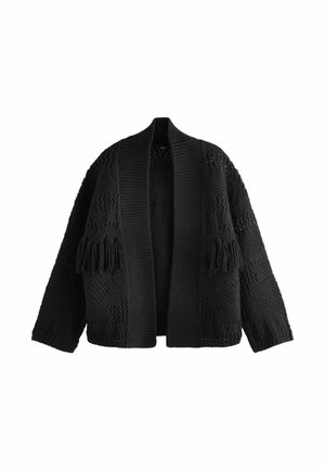 Femme portant un haut blanc côtelé sans manches, un cardigan noir en grosse maille et un pantalon à imprimé léopard avec des bagues argentées aux doigts.