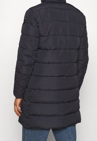 Marinblå lång puffjacka med ett kviltat mönster. Har huva och elastiska ärmslut. Tillverkad av lätt, vattenavvisande material.