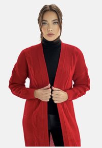 Röd stickad cardigan med ribbat mönster och flätstickade detaljer fram; öppen design med långa ärmar, bärs över en svart polotröja.