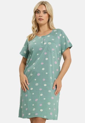 Frau mit blondem Haar, die ein lockeres, kurzärmeliges grünes Kleid mit einem Muster aus weißen und rosa Gänseblümchen trägt und vor einem einfarbigen Hintergrund steht.