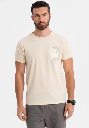 Beige katoenen t-shirt met een ronde hals en korte mouwen. De linkerkant bevat een borstzak met een licht camouflagedesign. Eenvoudig, casual ontwerp.
