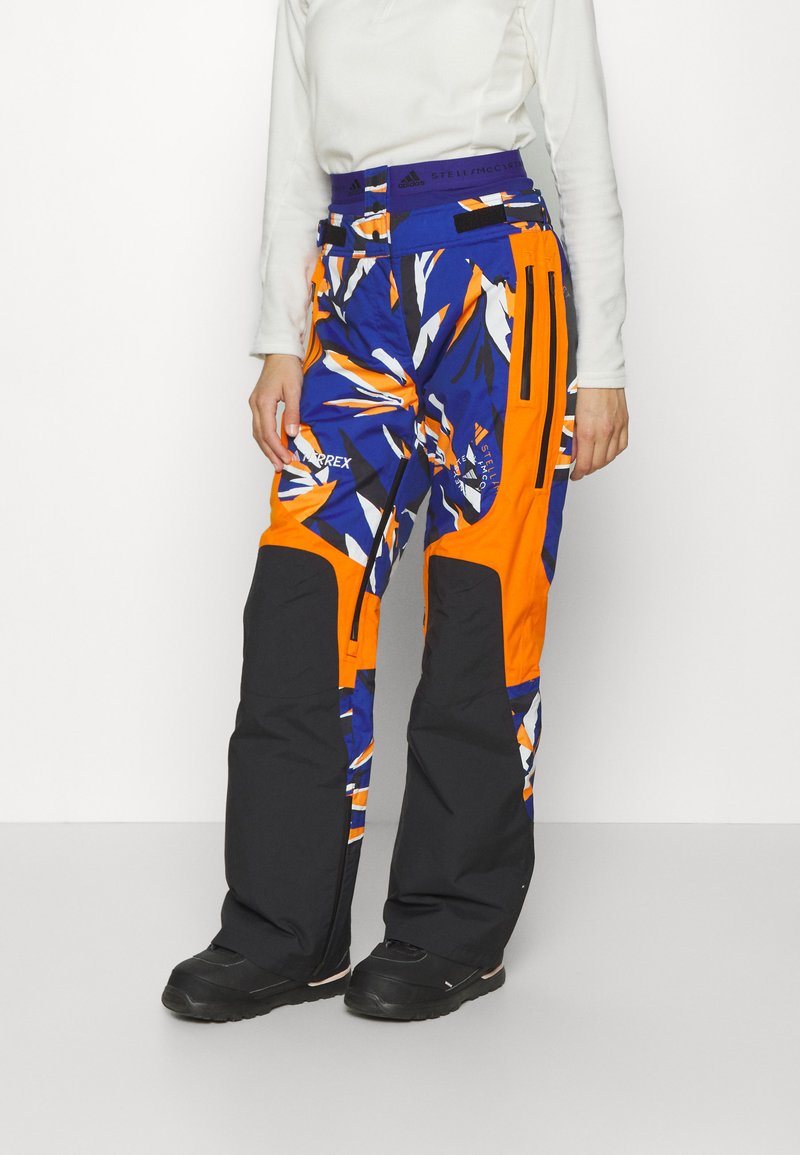 Persona che indossa pantaloni da esterno a motivo blu, arancione e nero con stivali neri, in piedi davanti a uno sfondo bianco semplice.