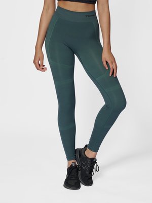Kvinde iført mørk teal seamless leggings med høj talje og sorte løbesko, står med det ene ben let bøjet mod en ensfarvet baggrund.