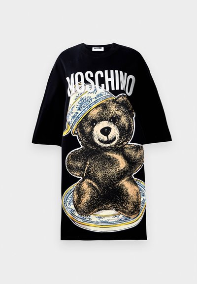 Melna oversize T-kreklā ar lielu attēlu, kurā ir lācis uz šķīvja ar zilu rakstainu apmali. Vārds "Moschino" drukāts treknās baltās burtos virs attēla.