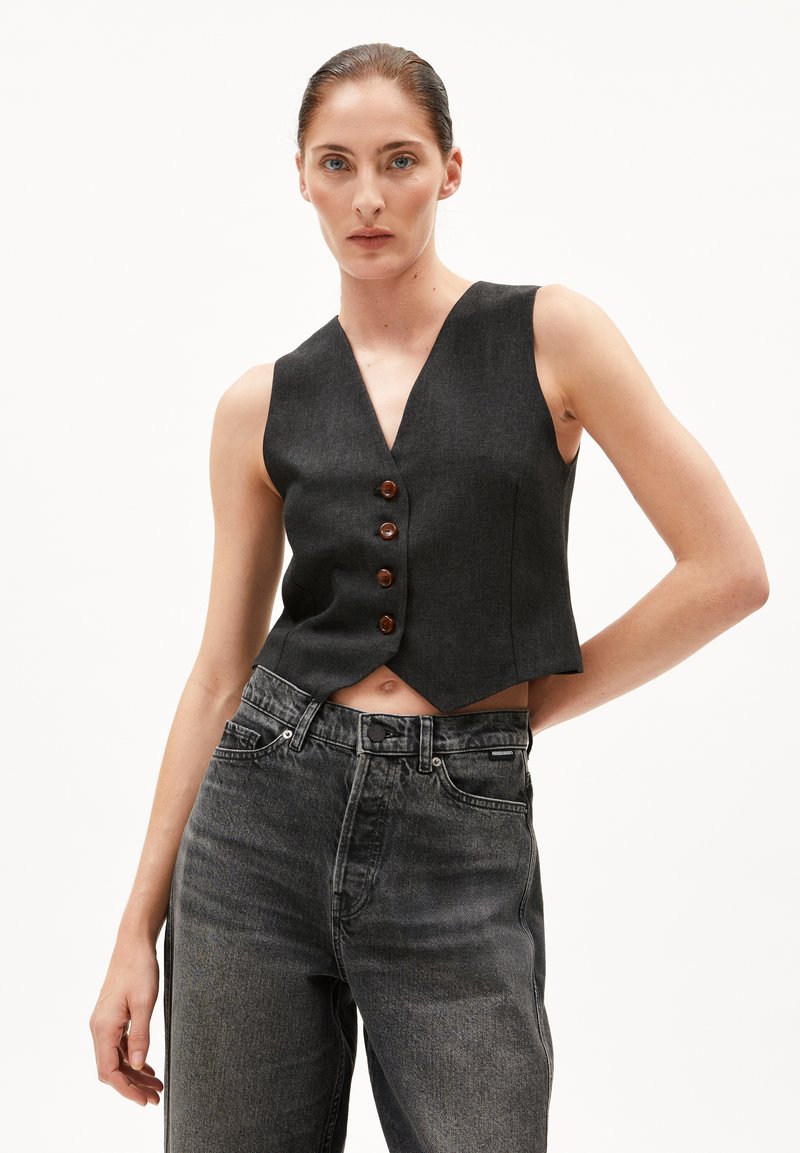 Dunkelblauer, verkürzter Jeansstapel mit V-Ausschnitt, der über fünf braune Knöpfe verfügt. Kombiniert mit hoch taillierten, leicht verwaschenen schwarzen Jeans.