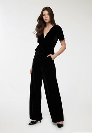 Jumpsuit in velluto nero con scollatura a V profonda, maniche corte e design a gamba larga. Presenta una vita annodata e tasche laterali. Texture liscia.