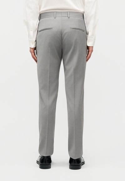 Άνδρας που φοράει ανοιχτό γκρι φόρμα παντελόνι, λευκό πουκάμισο και μαύρα παπούτσια, στέκεται γυρισμένος προς τα πίσω με φόντο ένα απλό υπόβαθρο.