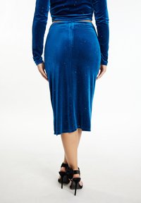 Blauwe fluwelen rok met een middellange ontwerp, voorzien van een subtiele glans en een split aan de achterkant. G gedragen met zwarte hoge hakken met een strikdetail.