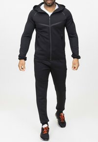 Haut de survêtement noir avec zip sur le devant, tissu texturé et capuche. Assorti à un pantalon de jogging et des baskets noires avec des accents orange.