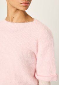 Lichtroze wollen truien met korte, opgerolde mouwen en een zachte, pluizige textuur. Wijde halslijn en subtiele ribbel-details aan de zoom.