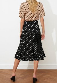 Schwarzer Rock mit weißen Punkten und ausgestelltem Saum, kombiniert mit einem beigen Oberteil mit weißen Punkten. Der Rock hat eine taillierte Passform und ein leichtes Rüschendetail. Glänzende schwarze Pumps.