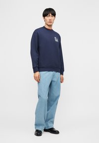 Joven con sudadera azul marino con el logo "EA", vaqueros de pierna ancha azul claro y zapatos negros, de pie contra un fondo blanco.