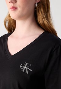 Melna V-veida T-krekls, izgatavots no kokvilnas, ar sudraba logo kreisā krūts daļā. Gluda tekstūra ar īsām, brīvām piedurknēm.