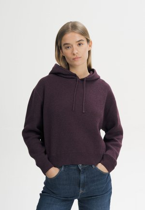 Jonge vrouw met stijl blond haar, een donkerpaarse hoodie en blauwe jeans, staand met handen in de zakken tegen een witte achtergrond.