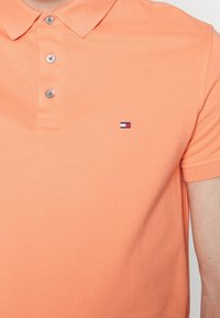 Polo tričko s krátkým rukávem v světle oranžové barvě. Má klasický límec, zapínání na tři knoflíky a malé vyšívané logo na prsou.