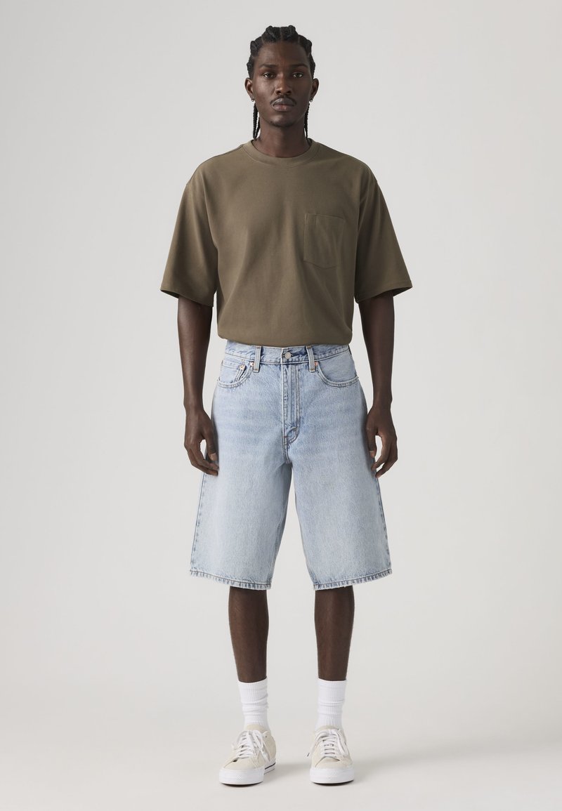 Mann mit geflochtenem Haar, der ein olivgrünes, übergroßes T-Shirt, hellblaue, knielange Jeansshorts, weiße Socken und beige Turnschuhe trägt.