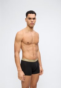 Μυώδης άνδρας με κοντά σγουρά μαλλιά, όρθιος, φοράει μαύρα σλιπ Tommy Hilfiger με μπεζ ζώνη μέσης, ουδέτερο γκρι φόντο.