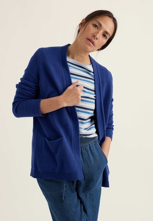 Žena v modrém kardiganu, pruhovaném modro-bílém tričku a volných džínových kalhotách pózující s jednou rukou v kapse a druhou držící kardigan.